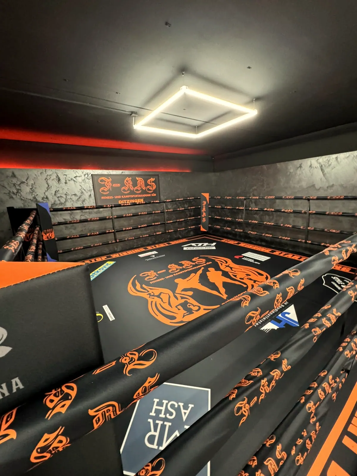 Boxring mit orange bedruckter Ringplane und Seilen unter einer LED-Decke